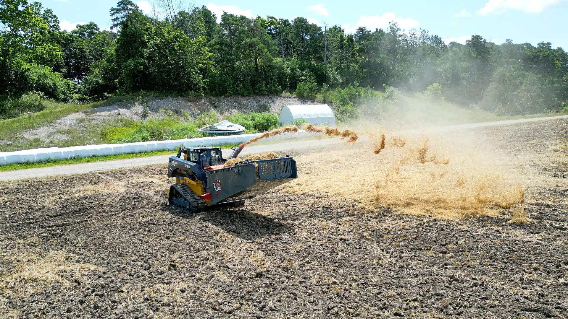 a John Deere skid steer with a straw bale blower is blowing straw in a large field for landscaping and farming purposes