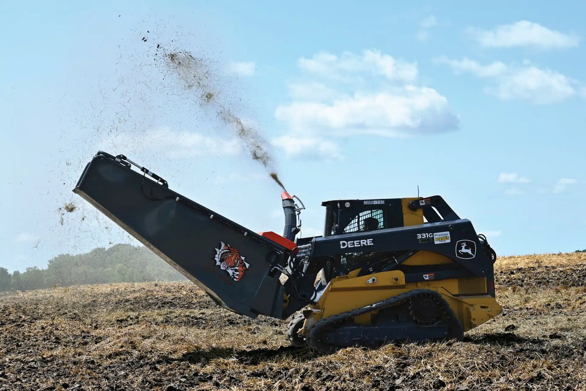 a John Deere skid steer with a straw bale blower is blowing straw in a large field for landscaping and farming purposes