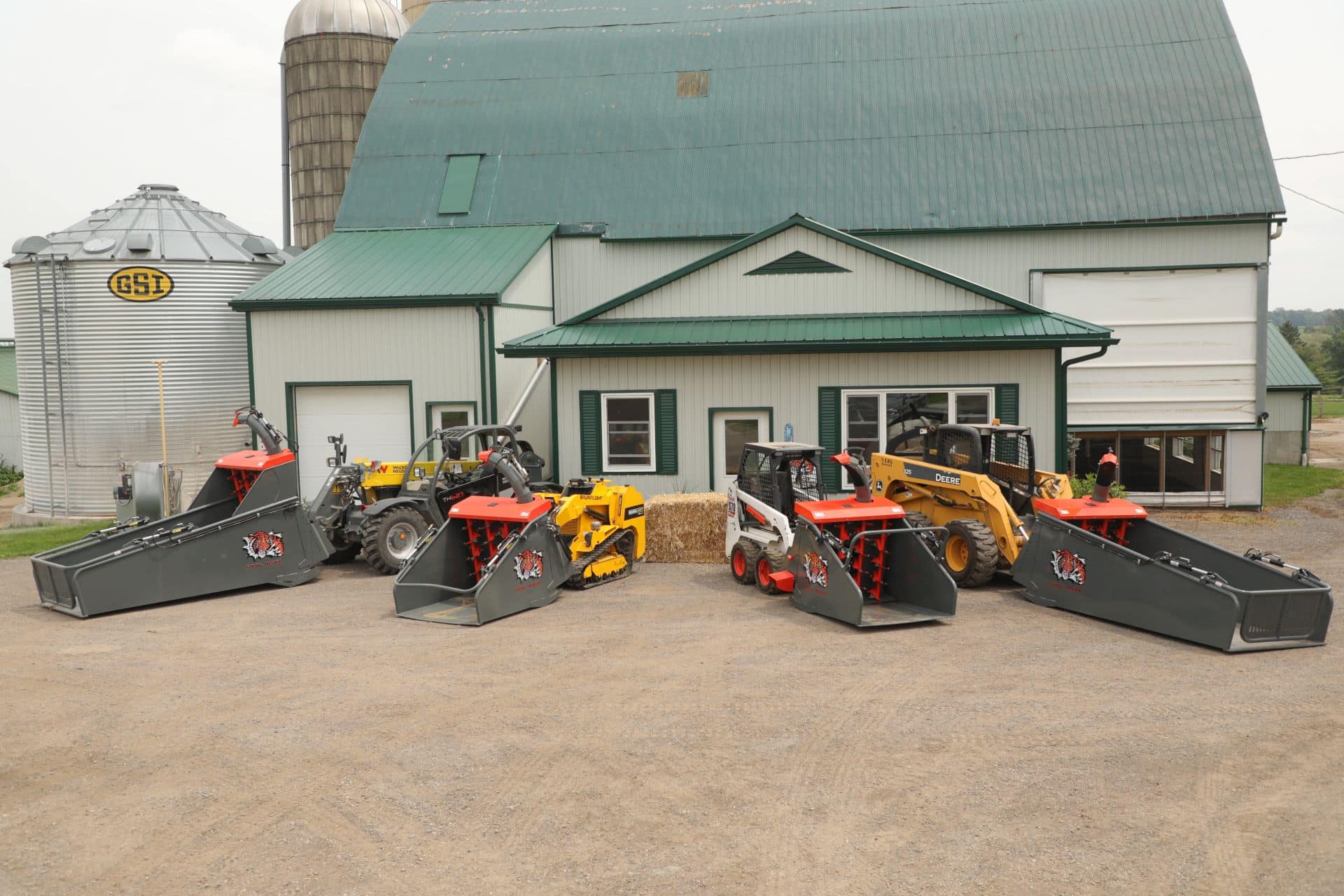a variety of skids steers, each with a different straw blower attached to them, sit in front of a barn