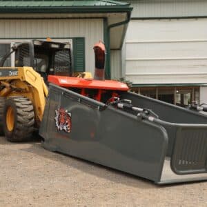 A John Deere skid steer has a large Strawmaster straw processor attached to it in front of a barn