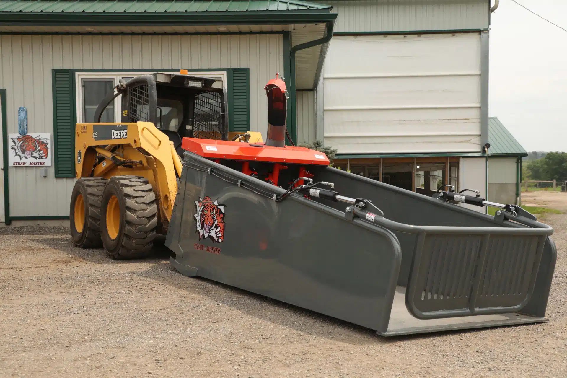 A John Deere skid steer has a large Strawmaster straw processor attached to it in front of a barn